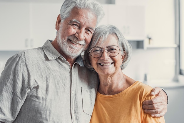 Photo headshot portrait of smiling elderly 60s husband and wife sit relax on couch hugging cuddling happy mature old couple rest on sofa in living room embrace look at camera show love and care