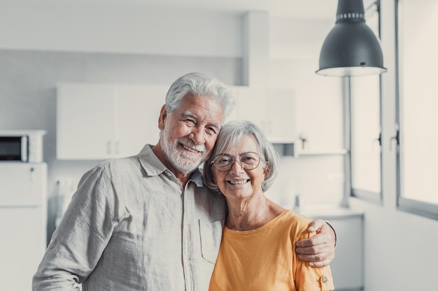 笑顔の60代の高齢者の夫と妻が立っているヘッドショットの肖像画がリラックスして抱き合い、幸せな成熟した老夫婦がリビングルームで休み、抱き合い、カメラを見て、愛とケアを示す