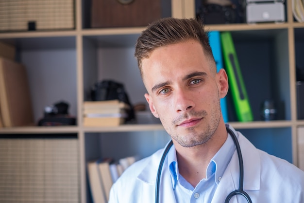 Headshot portrait of smiling young male GP or physician in white medical uniform and glasses look at camera posing in hospital, happy Caucasian man doctor show confidence skills in clinic workplace