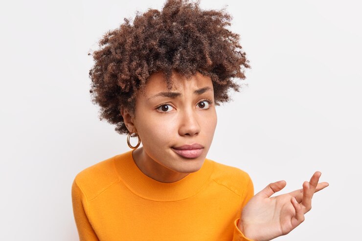 Headshot of puzzled African American woman looks with indignant expressio  listens attentively and tries to prove her case has curly hair wears casual jumper poses indoor against white wall
