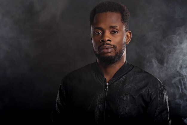 Photo headshot of serious dark skinned male holding chin looking mysteriously at camera, dressed in black windbreaker jacket, wristwatces, standing against dark background with white smoke. close up.