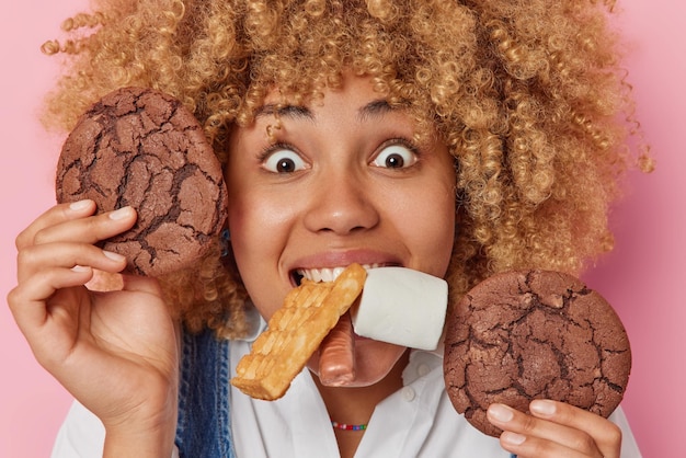 Headshot of surprised curly haired young woman eats delicious cookie waffle candy and marshmallow has sweet tooth addicted to sugar enjoys tasty snack poses indoor Unhealthy nutrition diet