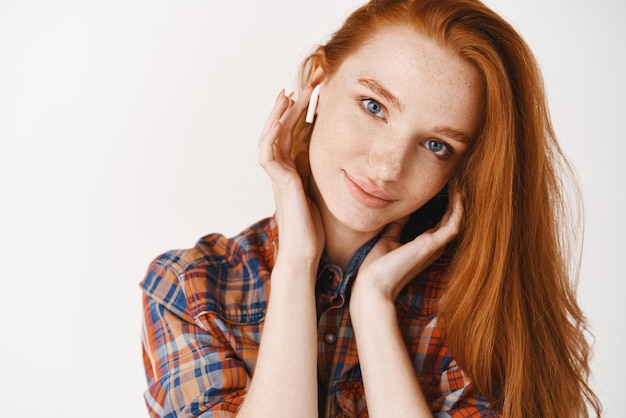 Foto headshot van aantrekkelijke jonge vrouw met lang rood haar en blauwe ogen die muziek luistert in draadloze hoofdtelefoons die dromerig glimlachen naar camera witte achtergrond