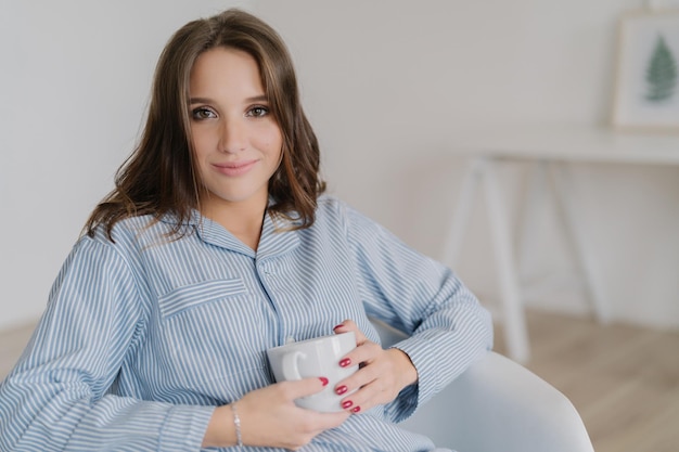 Headshot van blanke vrouw met make-up gekleed in pyjama houdt mok thee zit in comfortabele fauteuil geniet van goede rust thuis kijkt met aantrekkelijke uitdrukking op camera Binnenlandse sfeer