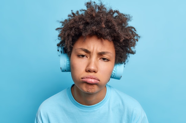 Headshot van gefrustreerde Afro-Amerikaanse vrouw met krullend haar portemonnees lippen maakt ongelukkige grimas luistert audiotrack in koptelefoon moe na een wandeling geïsoleerd over blauwe muur. slechte emoties