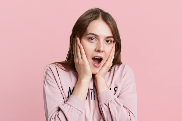 Headshot van geschokte mooie vrouw houdt handen op de wangen, draagt casual trui, geïsoleerd over roze muur. Adorable vrouw drukt grote verbazing uit iets te horen