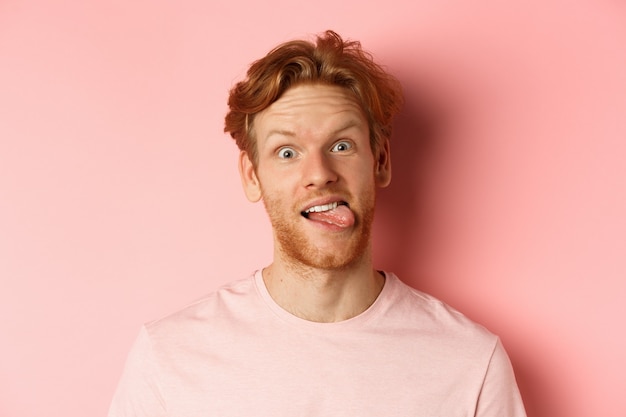 Headshot van grappige roodharige kerel die tong toont, domme gezichten naar de camera trekt, vrolijk staand tegen een roze achtergrond.