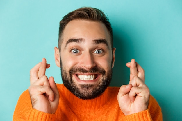 Headshot van hoopvolle bebaarde man die een wens doet, glimlachend en houdt vingers gekruist voor geluk, staande over lichtblauwe achtergrond.