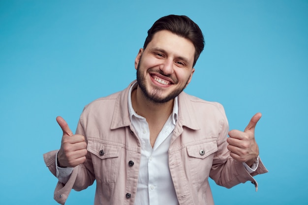 Headshot van jonge man draagt roze stijlvolle jeans jasje glimlachend en duim opdagen over blauwe achtergrond