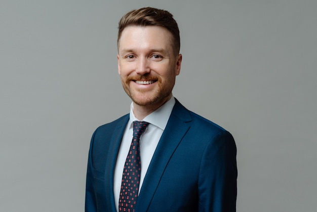 Headshot of young happy successful male office worker businessman wearing suit smiling at camera with positive face expression while posing against grey studio background. Business people concept