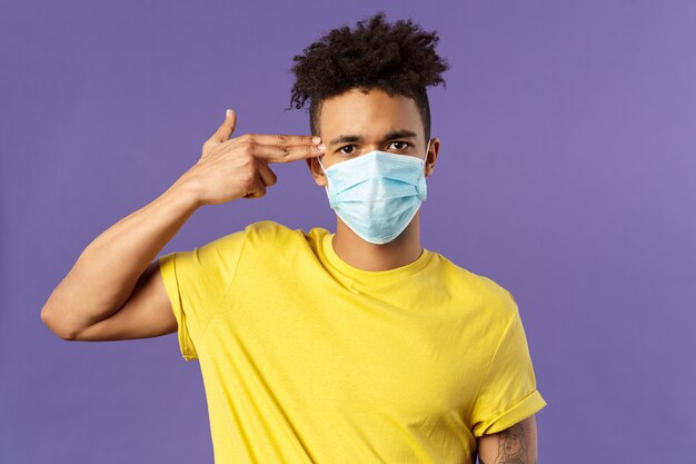 healtcare and medicine concept. Close-up portrait of serious hispanic guy in medical mask, showing gun near head as if sick and tired of people not avoid social contact, purple background