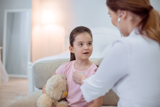 Health assessment. Focused beautiful girl sitting on blurred background and opening mouth while female doctor using stethoscope