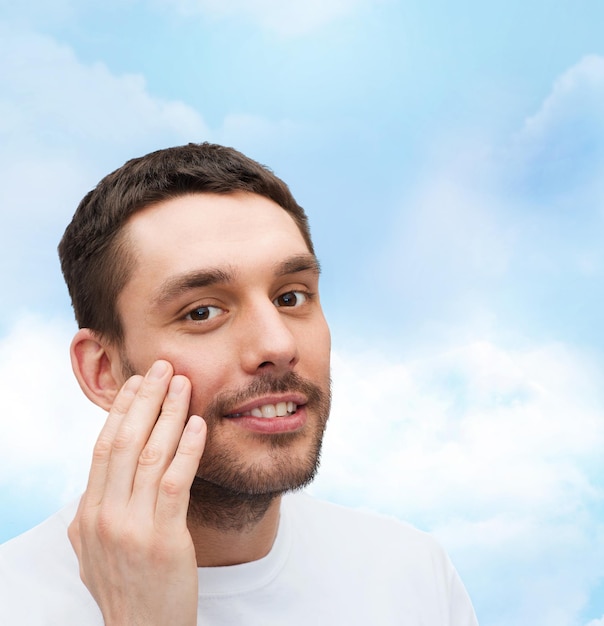 health and beauty concept - beautiful smiling man touching his face or applying cream