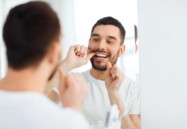 health care, dental hygiene, people and beauty concept - smiling young man with floss cleaning teeth and looking to mirror at home bathroom