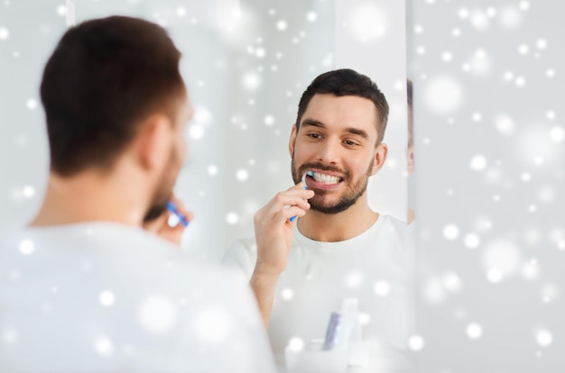 건강 관리, 치과 위생, 사람 및 미용 개념 - 칫솔로 치아를 닦고 눈 위에 집 화장실에서 거울을 보고 웃는 청년
