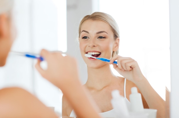 health care, dental hygiene, people and beauty concept - smiling young woman with toothbrush cleaning teeth and looking to mirror at home bathroom