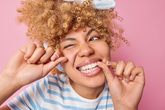 Health care dentistry and oral hygiene concept curly haired\
young woman uses dental floss to prevent oral disease winks eye\
looks away poses against pink background daily routines caries\
prevention