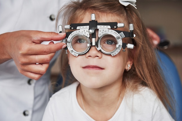 目の健康管理。医者は小さな女の子の視力をチェックし、フォロプターを調整します。