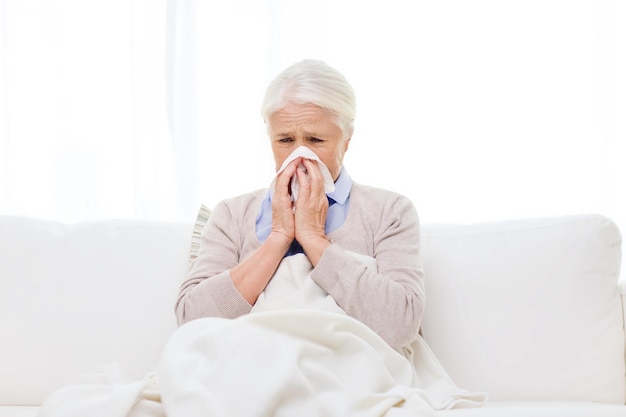 health care, flu, hygiene, age and people concept - sick senior woman blowing nose to paper napkin at home