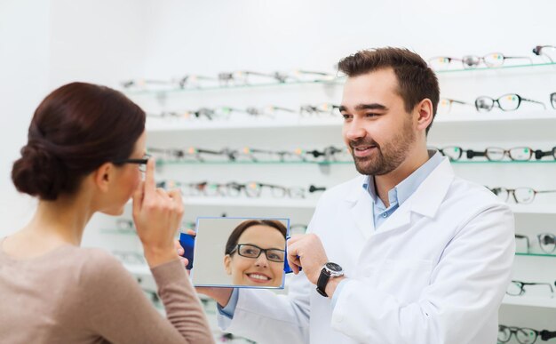 health care, people, eyesight and vision concept - optician and woman in glasses looking to mirror at optics store