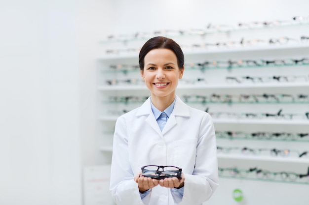 health care, people, eyesight and vision concept - smiling woman optician holding glasses at optics store