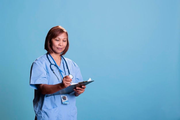 Photo health care worker wearing stethoscope smiling at camera while writing medical treatment on clipboard during clinical consultation. asian nurse checking patient health insurance, medicine concept