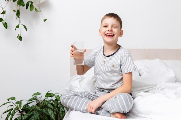 건강 개념 행복한 활동적인 아이는 아침에 침대에 있는 유리잔에 신선한 순수한 물을 들고 웃으면서 몸을 돌봅니다.