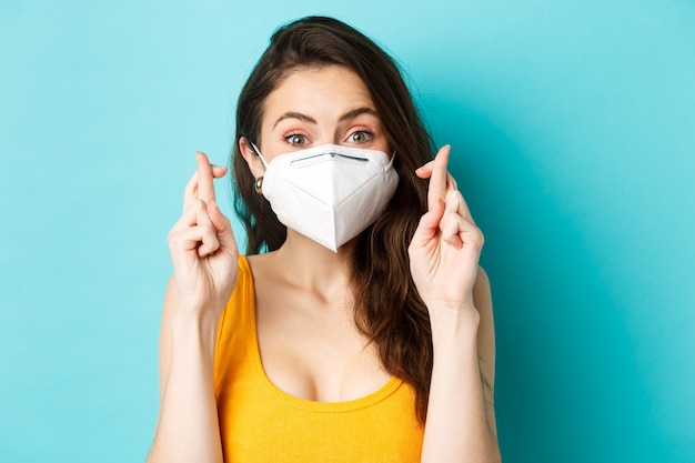 Health covid and lockdown concept close up of hopeful young woman in respirator mask cross fingers a...