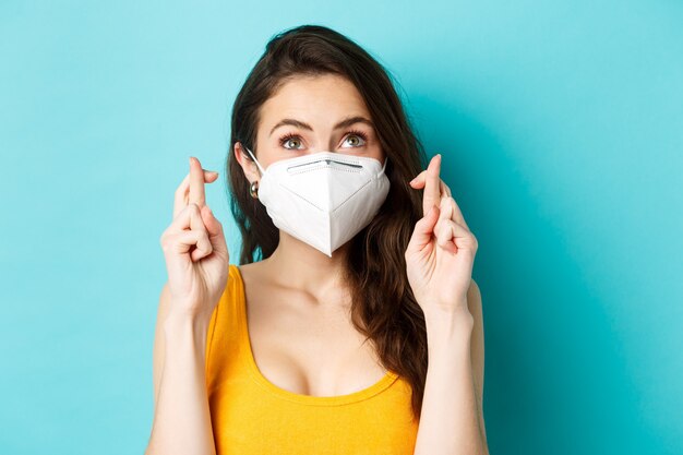 Health covid and lockdown concept close up of hopeful young woman in respirator mask cross fingers a...