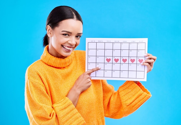 Health portrait and female with a calendar in a studio to track her menstrual or ovulation cycle Happy smile and face of a woman model pointing to a paper period chart isolated by blue background