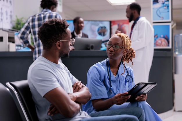 Health specialist doing consultation with patient, taking notes on digital tablet and writing report in waiting room. Medical assistant talking to man, consulting person with disease in waiting area.