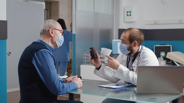 Health specialist giving bottle of pills at treatment to retired patient with face mask, helping senior man with disease diagnosis. Doctor showing drugs medication to elder person.