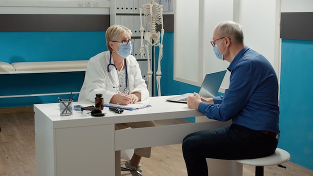 Health specialist taking notes at consultation appointment with old man during coronavirus pandemic. Writing checkup visit report on documents to give prescrption treatment in cabinet.