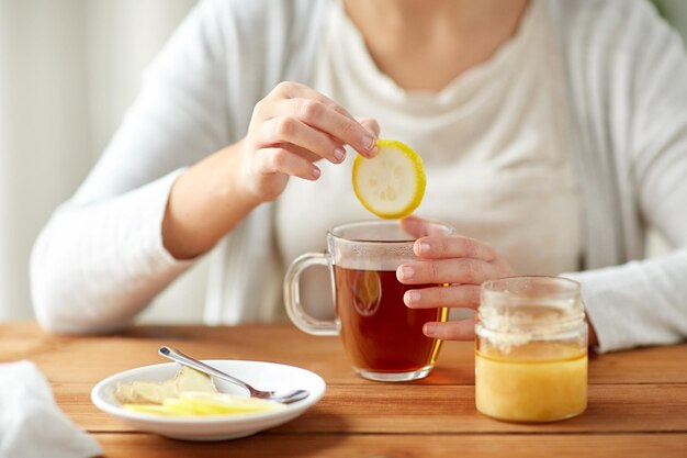 Photo health, traditional medicine and ethnoscience concept - close up of ill woman drinking tea with lemon, honey and ginger at wooden table