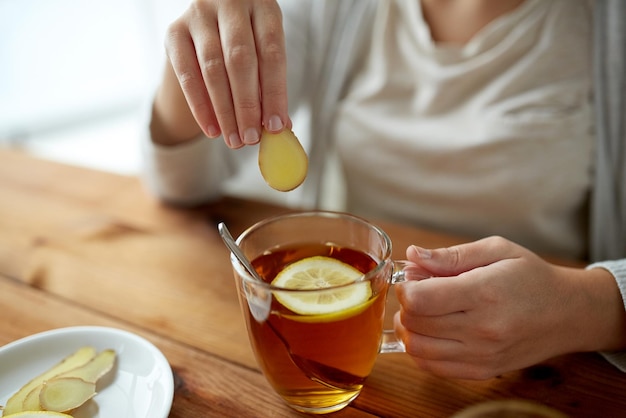 health, traditional medicine and ethnoscience concept - close up of woman adding ginger to tea cup with lemon