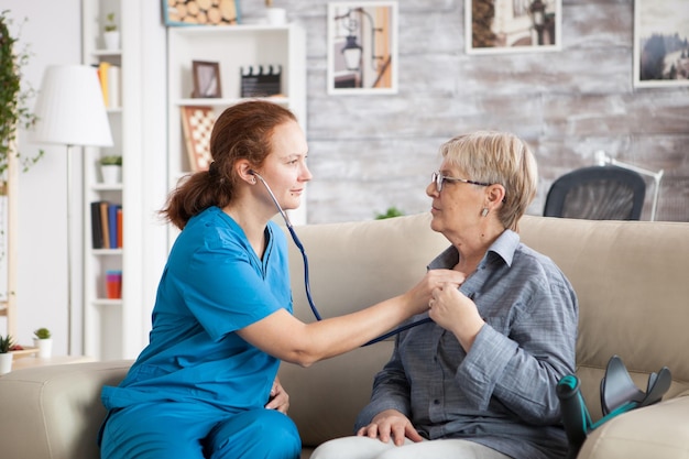 Health visitor and a senior woman in nursing home sitting on couch. Nurse checking old woman heartbeat with stethoscope.