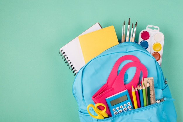 Healthcare concept. Studies during quarantine concept. Top above overhead view photo of backpack filled with colorful stationery and mask isolated on turquoise background