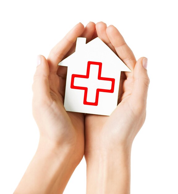 healthcare, medicine and charity concept - hands holding white paper house with red cross sign