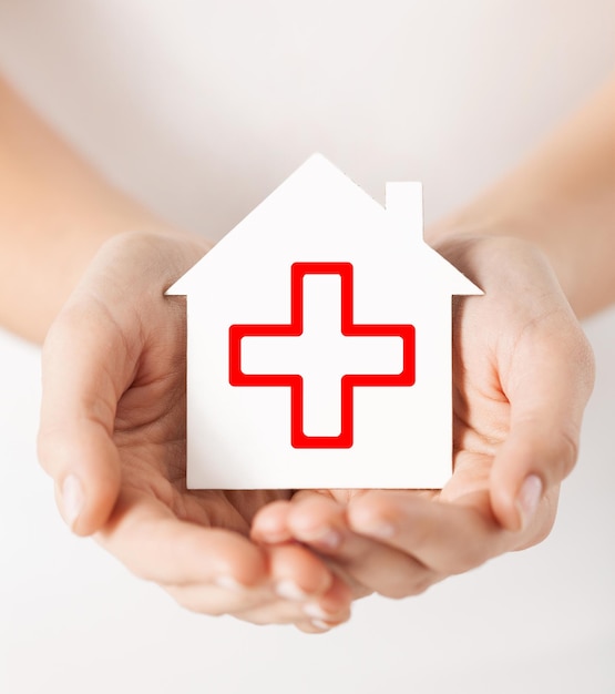 healthcare, medicine and charity concept - hands holding white paper house with red cross sign