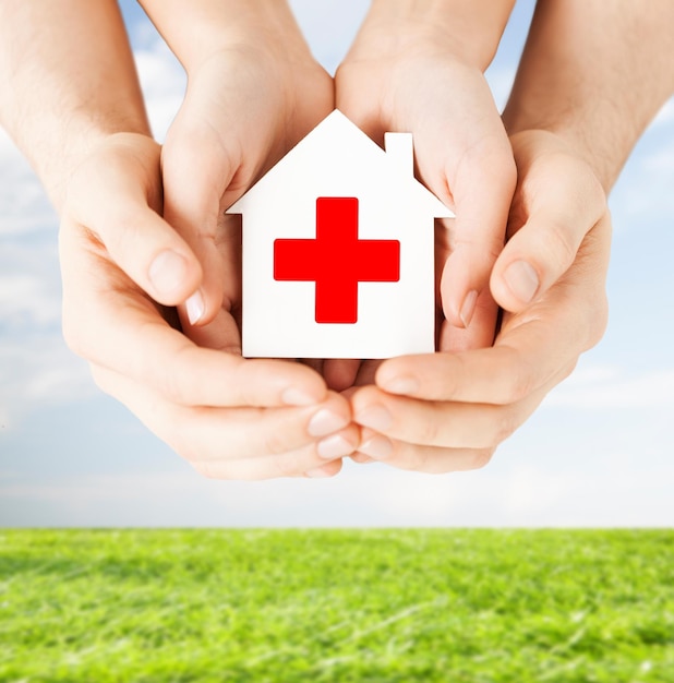 healthcare, medicine and charity concept - male and female hands holding white paper house with red cross sign