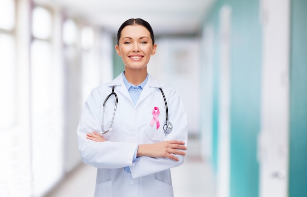 healthcare and medicine concept - smiling female doctor with stethoscope and pink cancer awareness ribbon over hospital background