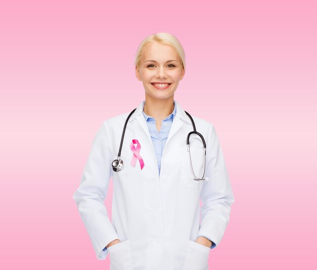 ヘルスケアと医学の概念-ピンクの背景の上に聴診器とピンクの癌意識リボンで笑顔の女性医師