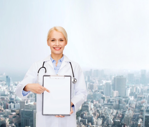 healthcare and medicine concept - smiling female doctor with stethoscope pointing finger to clipboard and blank page