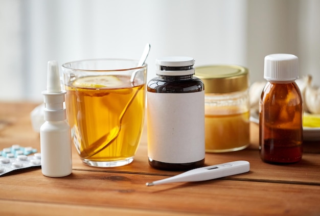 healthcare, medicine and treatment concept - drugs, thermometer, honey and cup of tea on wooden table