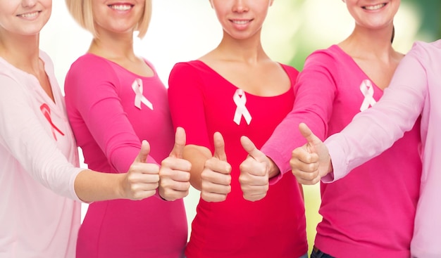 写真 ヘルスケア、人々、ジェスチャー、医学の概念-緑の背景の上に親指を示すピンクの乳がん啓発リボンと空白のシャツを着た笑顔の女性のクローズアップ