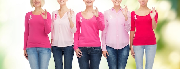 Foto concetto di assistenza sanitaria, persone, gesto e medicina - primo piano di donne sorridenti in camicie bianche con nastri rosa per la consapevolezza del cancro al seno che mostrano segno ok su sfondo verde