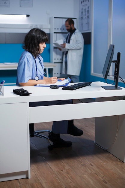 医療従事者はコンピュータで患者の保険をチェックし,メモを取ります 現代の診療所でデスクトップPCを使用して医療情報をチェックして比較する女性医師
