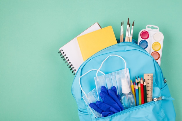 Healthcare at school concept. Top above overhead view photo of backpack filled with colorful stationery two blue masks rubber gloves and hand sanitizer isolated on turquoise background