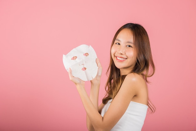 Healthcare spa. Asian woman holding purifying mask face on hands, Portrait beautiful female smiling holds facial sheet mask, Studio shot isolated on blue background, Face care and beauty treatments