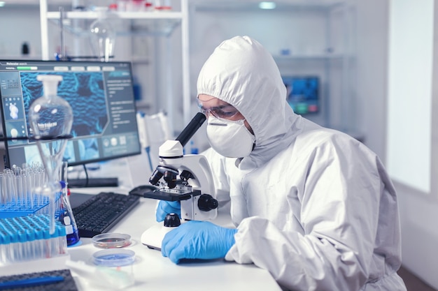 Healthcare specialist analyzing vaccine on microscope dressed in protection suit. Virolog in coverall during coronavirus outbreak conducting healthcare scientific analysis.